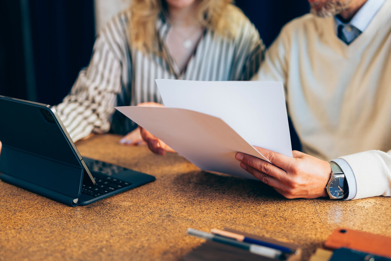 Two people sitting at a table reviewing documents, with one person holding printed papers and the other using a tablet with a keyboard case. Only their torsos and hands are visible. Office supplies are on the table.