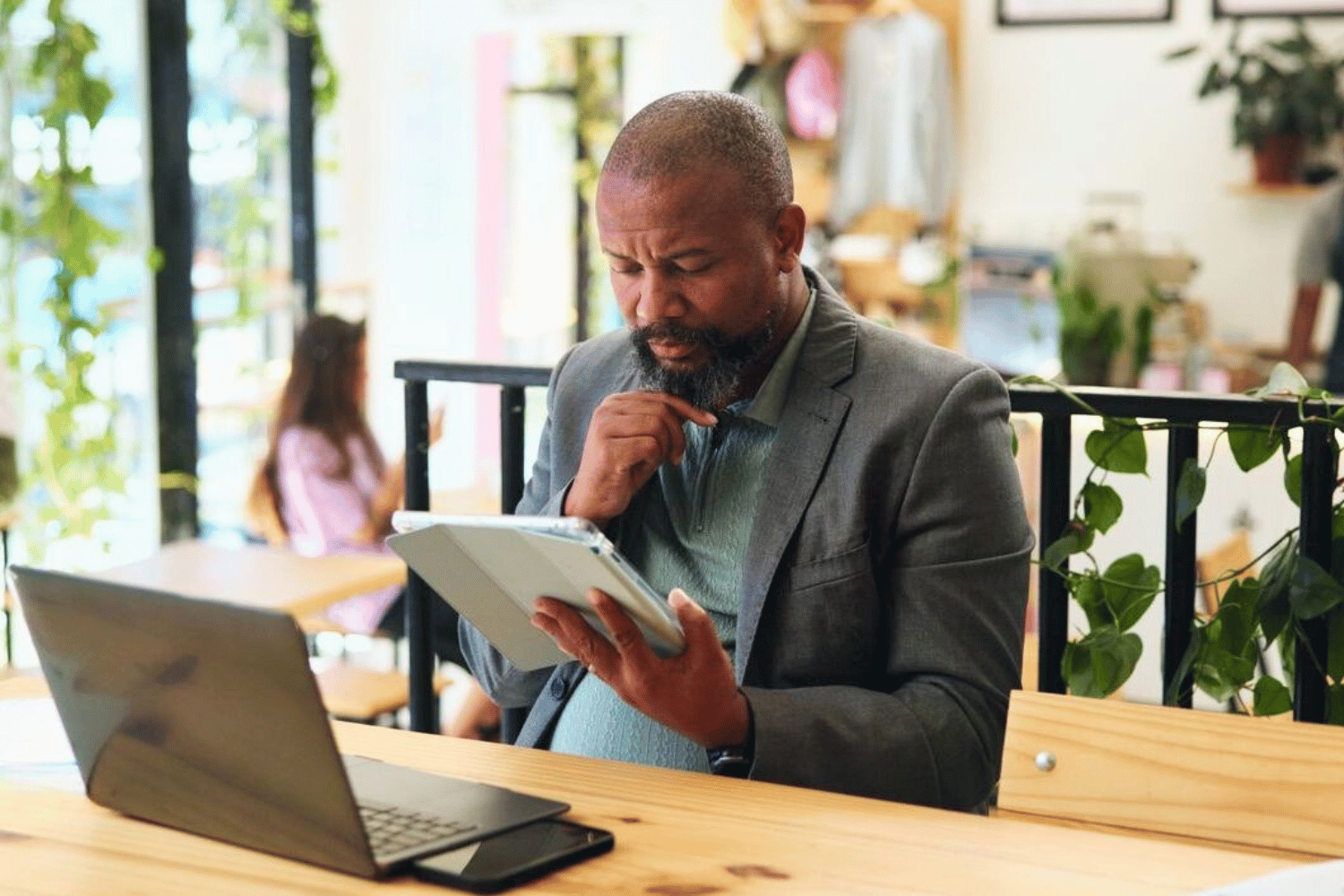 A man in a grey suit sits at a wooden table in a café, looking thoughtfully at a tablet in one hand while resting his other hand on his chin.