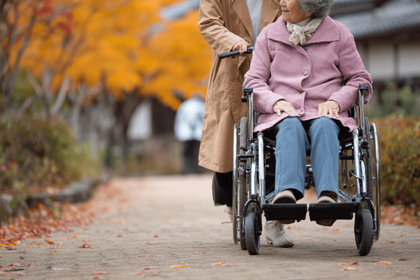An elderly woman in a wheelchair is being pushed by a caregiver along a path lined with autumn leaves and trees with vibrant orange foliage. The elderly woman is wearing a pink coat and looking back at the caregiver, who is dressed in a beige coat.