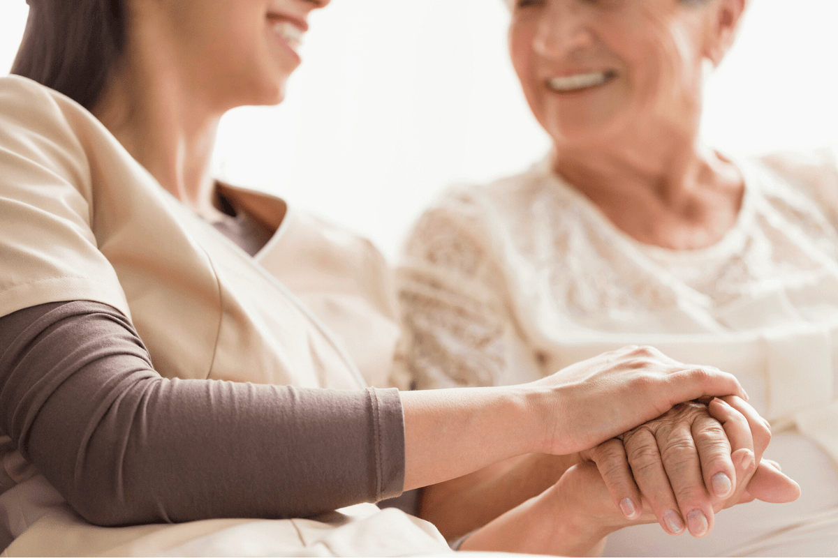A young caregiver and an older woman are sitting close together, smiling. The caregiver is gently holding the older woman's hand in both of hers, conveying warmth and support.