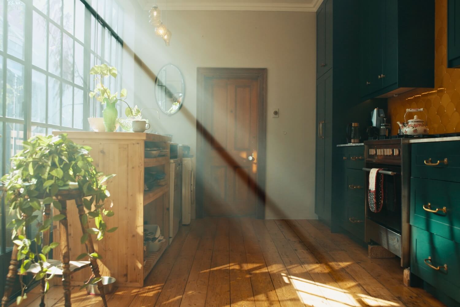 Sunlight streaming through large windows into a quiet kitchen with warm wood floors and green cabinets, evoking the stillness of a home after unexpected loss