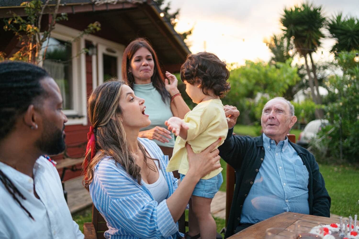 Several generations of a family gathered outdoors around a table; an older man smiles and reaches toward a toddler being held up by a woman, while other adults watch and smile in a garden setting near a house.