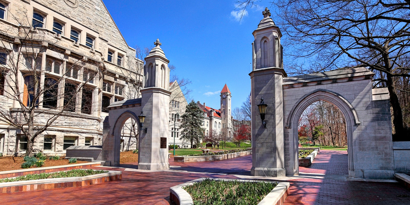 A bright, clear day at the Indiana University campus in Bloomington, Indiana. The iconic Sample Gates, made of light gray limestone, frame a red-brick pathway that leads into the university grounds. Historic campus buildings with pointed red roofs and stone facades are visible in the background, surrounded by trees just beginning to bloom.