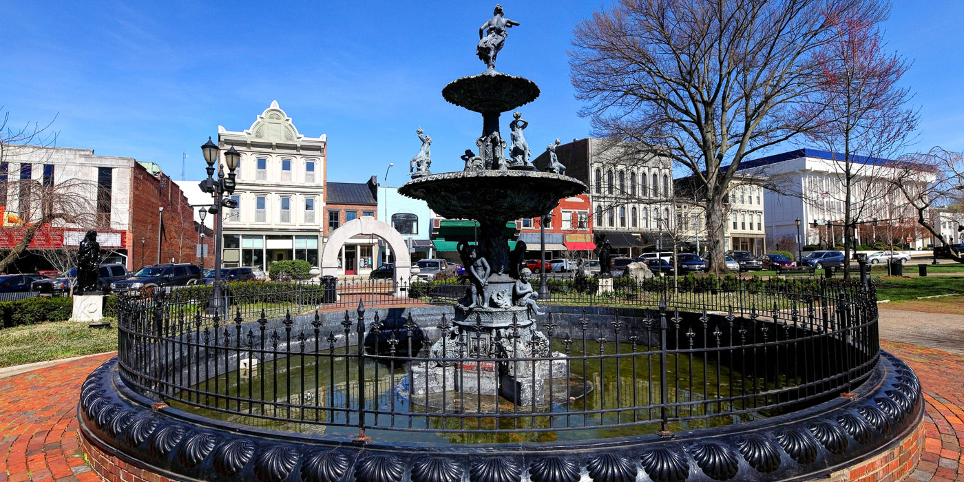 A historic cast-iron fountain surrounded by a decorative black fence stands in the center of Fountain Square Park in Bowling Green, Kentucky. The fountain features multiple tiers with sculpted figures, and water flows into a circular basin. In the background are colorful storefronts and buildings with classic architectural details, as well as trees and parked cars lining the street.