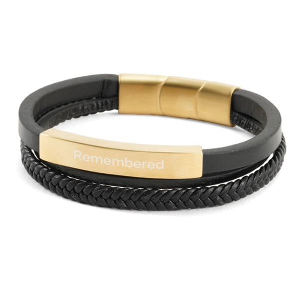 The Brass/Black Triple Band Leather Bracelet, available at Magnolia Cremations, is a contemporary memorial piece featuring three black leather bands—one smooth, one braided, and one accented with a gold-tone bar. The bar is engraved, showcasing the option for personalization. The bracelet secures with a gold-tone stainless steel clasp and discreetly holds a small portion of ashes, offering a stylish and meaningful way to keep a loved one close.
