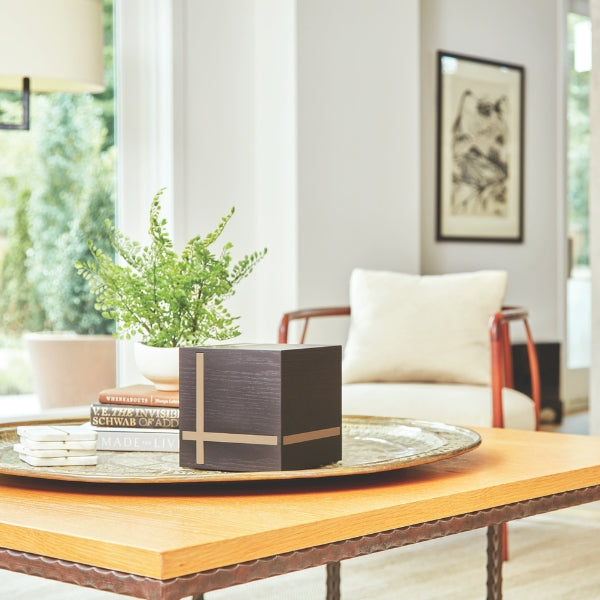 Bristol Ebony/Bamboo Extra-Large Cremation Urn displayed on a decorative tray in a modern living room. The urn features a sleek cube design with a deep ebony MDF finish and contrasting bamboo inlays in a geometric pattern. Designed for home display and can be personalized with engraving. Features a sliding bottom panel secured with a screw. 245 cubic inch capacity.