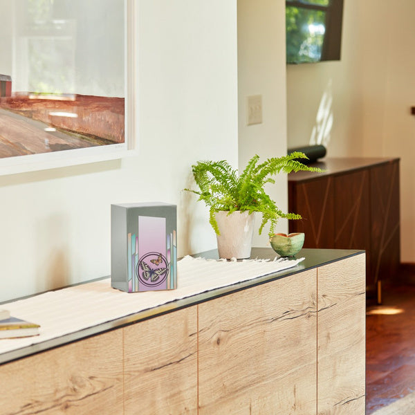 Life Canvas Butterflies Pewter Full-Size Cremation Urn displayed on a modern wooden console table in a well-lit home setting. The urn has a brushed silver stainless-steel finish with a soft pink and lavender gradient panel, two butterflies, and pastel geometric accents. Designed for home display and can be personalized with engraving. 200 cubic inch capacity.