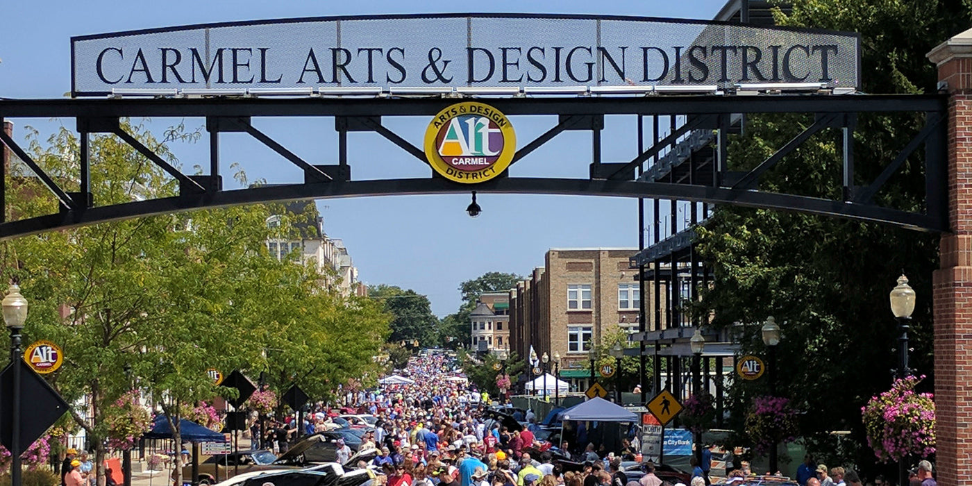 A bustling street scene in the Carmel Arts & Design District in Carmel, Indiana. A large overhead sign marks the entrance to the district, reading “Carmel Arts & Design District” with a colorful circular logo below it. The street is packed with people attending an outdoor event or festival, lined with green trees, hanging flower baskets, and a mix of modern and historic buildings on either side.