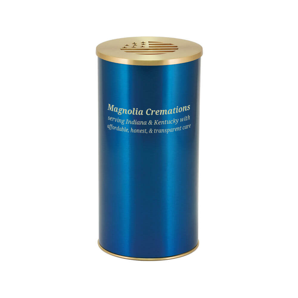A metallic blue cylindrical scattering urn with gold-colored lid and base. The lid features a cutout of a stylized American flag with stars and stripes. The front of the urn is labeled with the text: "Magnolia Cremations – serving Indiana & Kentucky with affordable, honest, & transparent care."