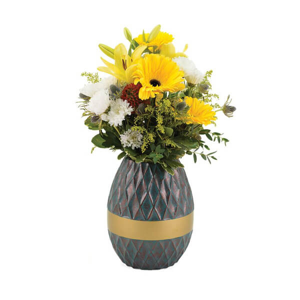 Decorative urn repurposed as a flower vase, holding a vibrant bouquet of yellow lilies, yellow gerbera daisies, white chrysanthemums, and assorted greenery. The urn has a textured, diamond-patterned teal and reddish surface with a wide gold band around the middle.