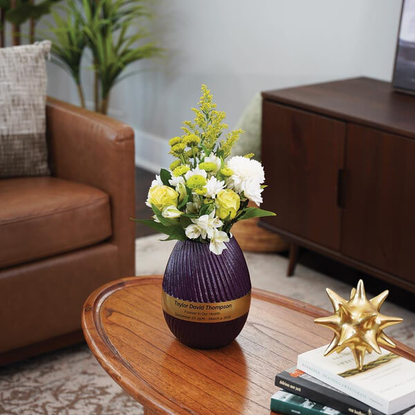 A purple, ribbed scattering urn with a gold band and engraved nameplate sits on a wooden coffee table, repurposed as a vase with a bouquet of white, yellow, and green flowers. The urn is placed in a modern living room with a leather chair, wooden cabinet, and decorative items.