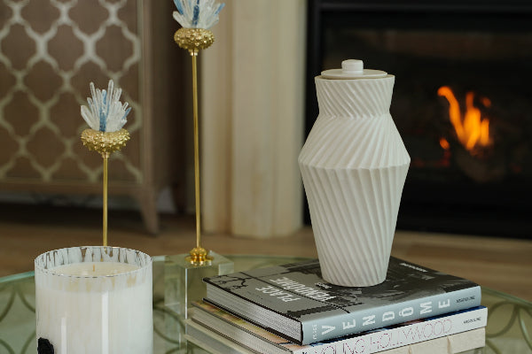 The Deco Twist Beach Ceramic Urn by Stoneware & Co., designed exclusively for Magnolia Cremations, displayed on a coffee table in a cozy living room setting. This handcrafted urn in tranquil beach beige features an Art Deco-inspired twisted groove pattern, creating depth and elegance. A timeless tribute, suitable for individuals weighing up to 210 lbs.