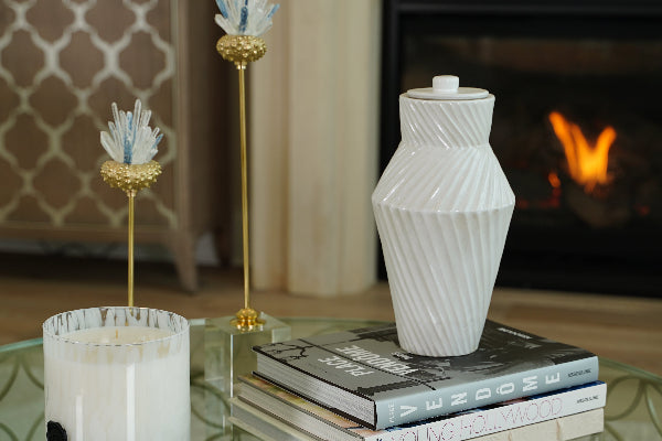 A Deco Twist Cloud Ceramic Urn by Stoneware & Co. is elegantly displayed atop a stack of books on a glass coffee table. The handcrafted urn features a calming cloud white finish with a unique twisted groove design, adding an Art Deco-inspired touch. A flickering fireplace in the background enhances the serene ambiance.