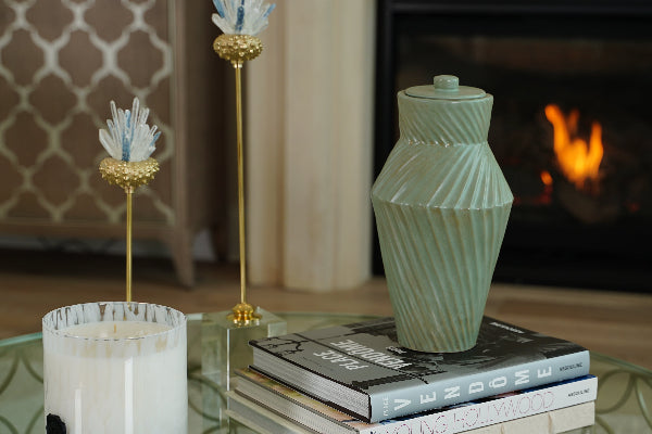 Deco Twist Forest Ceramic Urn by Stoneware & Co., displayed on a stack of books in front of a fireplace. The handcrafted urn features a deep forest green glaze with a striking twisted groove design, blending Art Deco elegance with a natural, earthy tone.