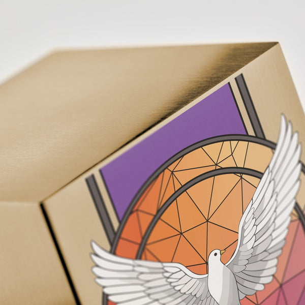 Close-up of a gold-colored urn showcasing a detailed illustration of a white dove with outstretched wings. The background features an intricate geometric stained glass-inspired design in shades of orange, red, and purple. The brushed metal surface of the urn is visible, reflecting light.