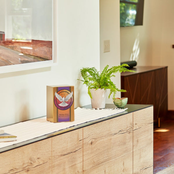 A gold-toned stainless steel urn featuring a white dove with outstretched wings against a stained glass-inspired background in warm orange and purple hues. The urn is displayed on a modern wooden console table, accompanied by a green potted fern and decorative elements, in a well-lit contemporary home setting.