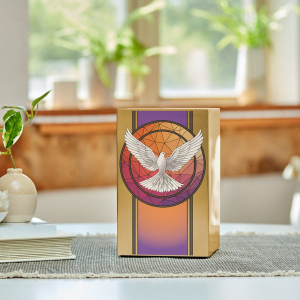A gold-colored stainless steel urn featuring a detailed white dove with outstretched wings, set against a geometric stained glass-inspired background in warm orange, purple, and red hues. The urn is placed on a table with a textured gray runner, accompanied by a small stack of books and a ceramic vase, with natural light streaming in from a nearby window.