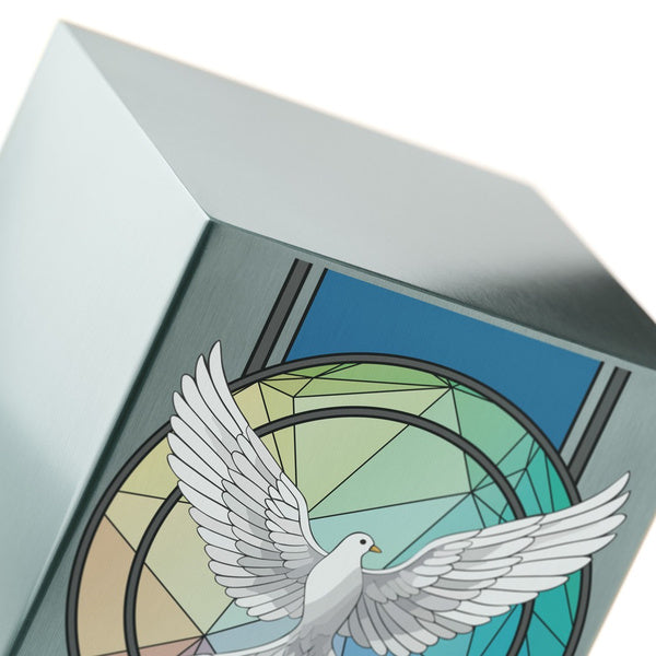 Close-up of a brushed silver stainless steel urn, highlighting its sharp edges and intricate dove design. The dove, with outstretched wings, is set against a stained glass-inspired background in shades of blue and green.