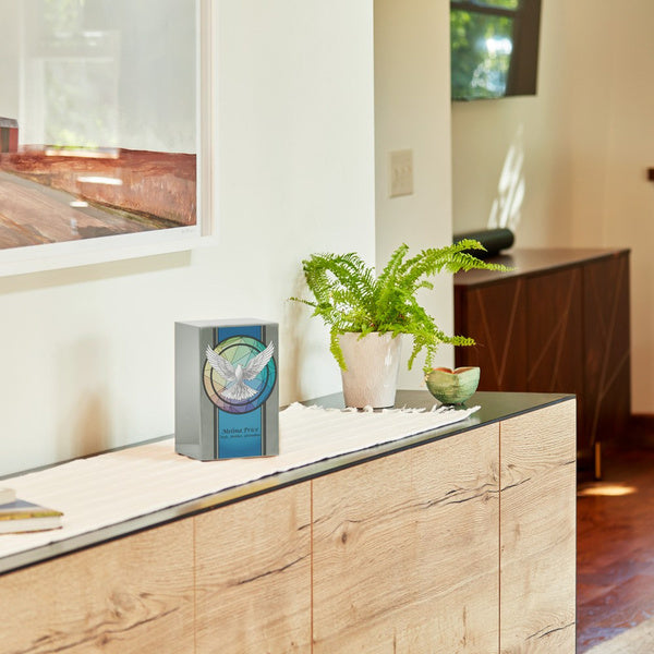 Stainless steel urn with a brushed pewter finish, featuring a colorful dove design, displayed on a modern wood console table. A green fern in a white pot sits nearby, with a framed artwork and a dark wood cabinet in the background.
