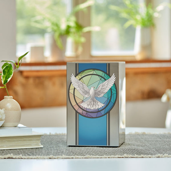 Brushed silver stainless steel urn featuring a vibrant dove design within a stained glass-inspired circle, placed on a woven mat with books and decorative accents nearby. Sunlight streams through a window, highlighting greenery in the background.