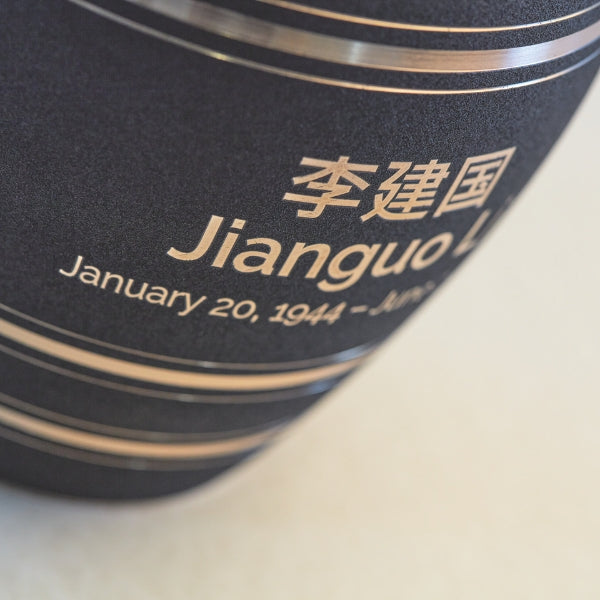 A close-up of a matte black memorial urn with gold accent stripes, showcasing an engraved name and dates in both Chinese characters and English. The engraving is clean and precise, contrasting against the dark background. Engraving is available for personalization.