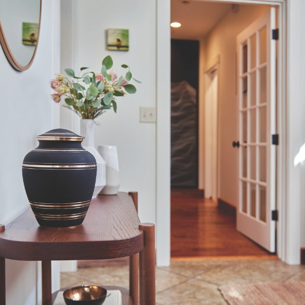 A matte black memorial urn with gold accent stripes sits on a wooden console table in a well-lit hallway. Behind the urn, a white vase with fresh flowers and two decorative ceramic vases add to the serene setting. A partially open French door and a warmly lit hallway are visible in the background.