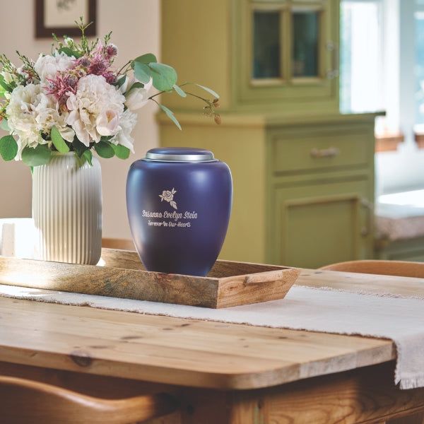 A deep purple memorial urn with a matte finish and a silver-toned lid, placed on a wooden tray atop a dining table. The urn features engraving, which is available for personalization. A vase with white and pink flowers sits beside it, with a rustic green cabinet in the background.