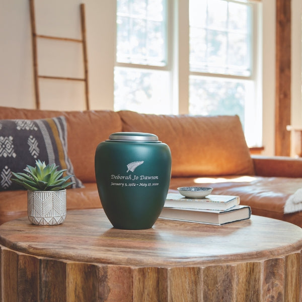 A matte emerald green memorial urn with a silver lid, placed on a rustic wooden coffee table in a warmly lit living room. The urn features an engraving, indicating the option for personalization. A small potted succulent and stacked books are nearby, with a brown leather couch and large windows in the background.