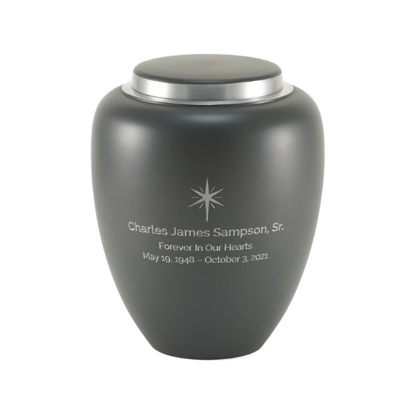 A matte granite gray memorial urn with a silver lid, featuring engraved text and a small starburst design on the front. The urn has a smooth, modern shape. Engraving is shown and available.