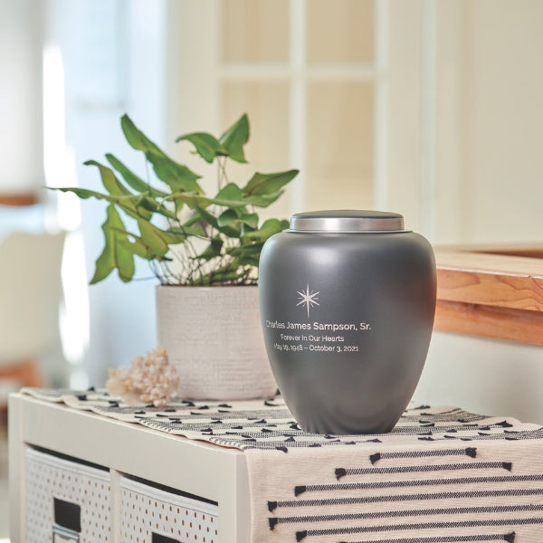 A matte granite gray memorial urn with a silver lid, placed on a patterned fabric atop a storage unit. The urn has engraved text on the front. A green potted plant and decorative elements are nearby, creating a peaceful setting. Engraving is shown and available.