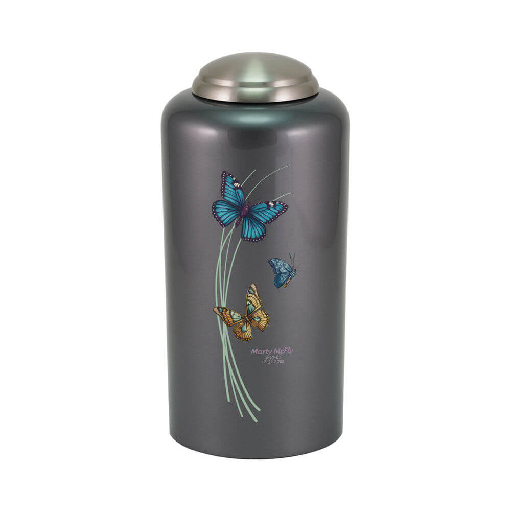 Dark gray cremation urn with a metallic finish and domed silver lid. The front features a design of three butterflies—blue, teal, and orange—fluttering around curved pale green lines. Below the design, optional engraved text displays a name and dates.