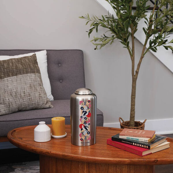 Silver cremation urn with a vertical Japanese-inspired design featuring cranes, red sun motifs, and floral patterns placed on a wooden coffee table in a modern living room. The table also holds a yellow candle, a small white jar, and a stack of books. A gray couch with cushions and a potted tree are in the background.