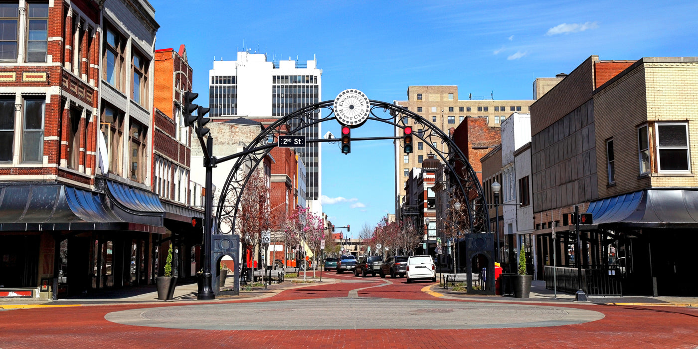A view of downtown Evansville, Indiana, showing a red-brick street lined with historic and modern buildings. An arched black metal sign spans across the street, reading "Evansville River City" above a traffic light. The street is relatively quiet, with a few parked cars and trees just starting to bloom with pink flowers under a bright blue sky.