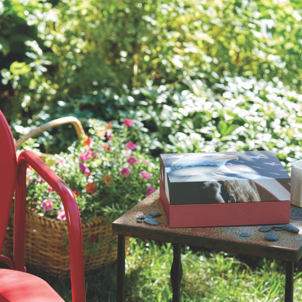 Biodegradable scattering urn with a patriotic American flag and eagle design sits on a small outdoor table surrounded by smooth stones. The urn has a red base and a patriotic-themed lid. In the background, a garden with green foliage and colorful flowers is visible, along with part of a red chair and a wicker basket.