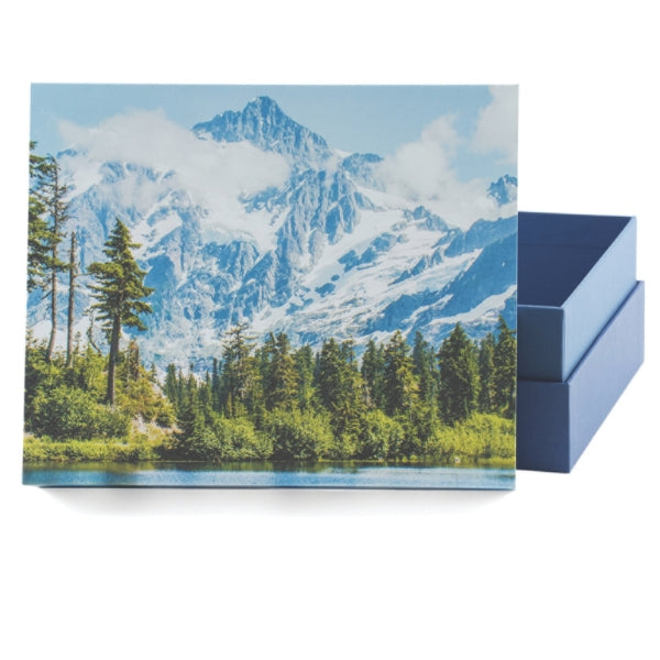 A biodegradable memorial urn with a removable lid, featuring a printed image of a snow-capped mountain, lush evergreen trees, and a calm lake. The urn is partially open, revealing its deep blue interior.