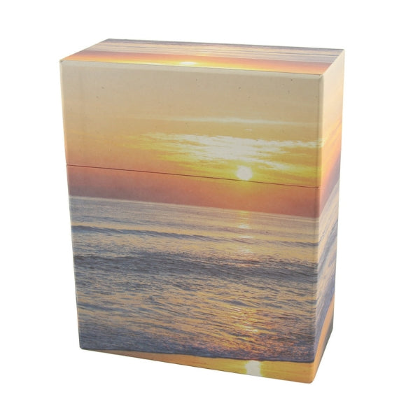 A biodegradable memorial urn with a printed image of a peaceful beach at sunset. The urn has a rectangular shape with a removable lid, designed for scattering or burial. The warm hues of the setting sun reflect on the ocean waves, creating a serene tribute.
