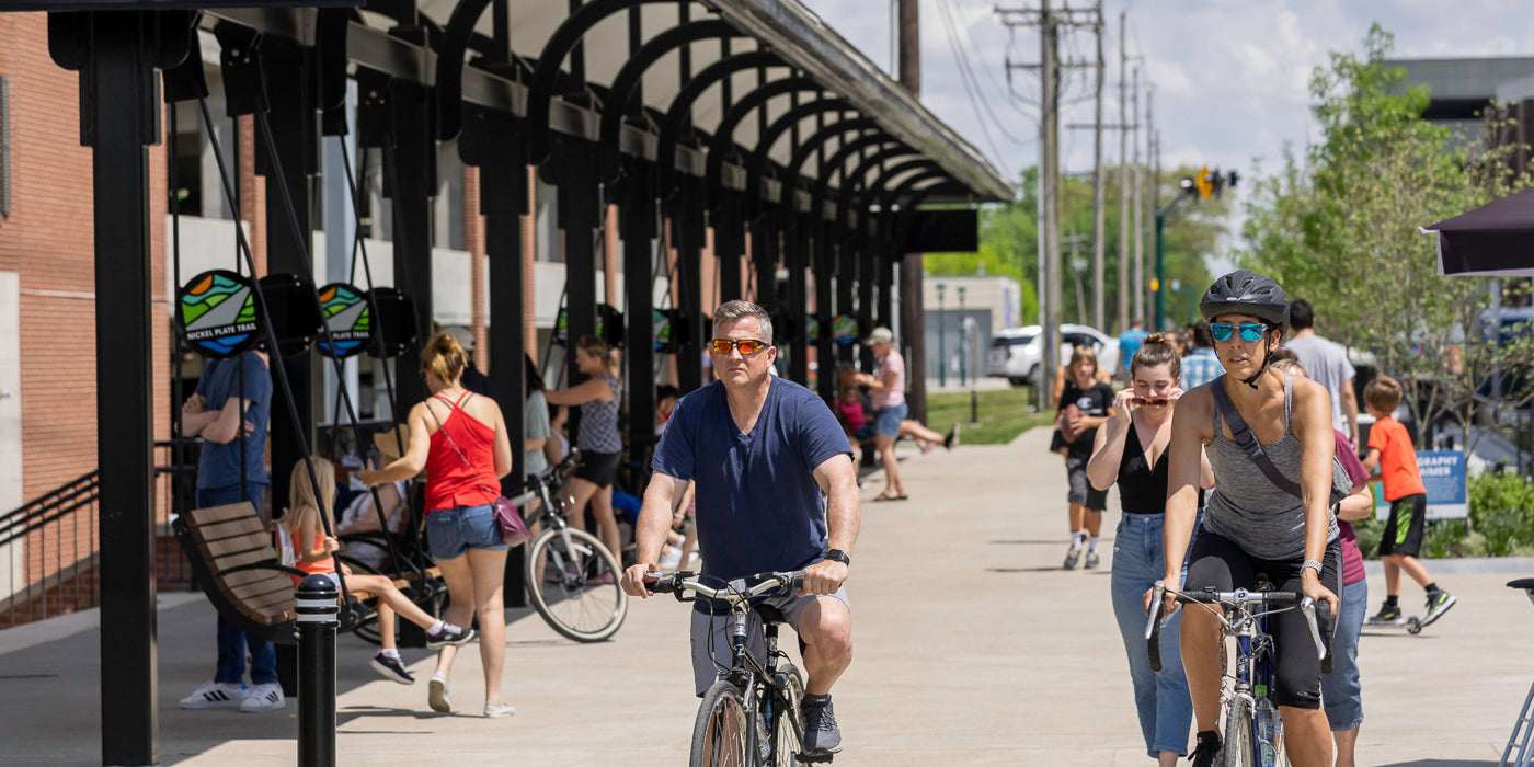 A lively scene along the Nickel Plate Trail in Fishers, Indiana. People are walking, biking, and relaxing on swing-style benches beneath a black metal canopy next to a brick building. A man and a woman in the foreground ride bicycles, with others walking and enjoying the sunny day in the background. The trail is busy and vibrant, lined with greenery and street activity.