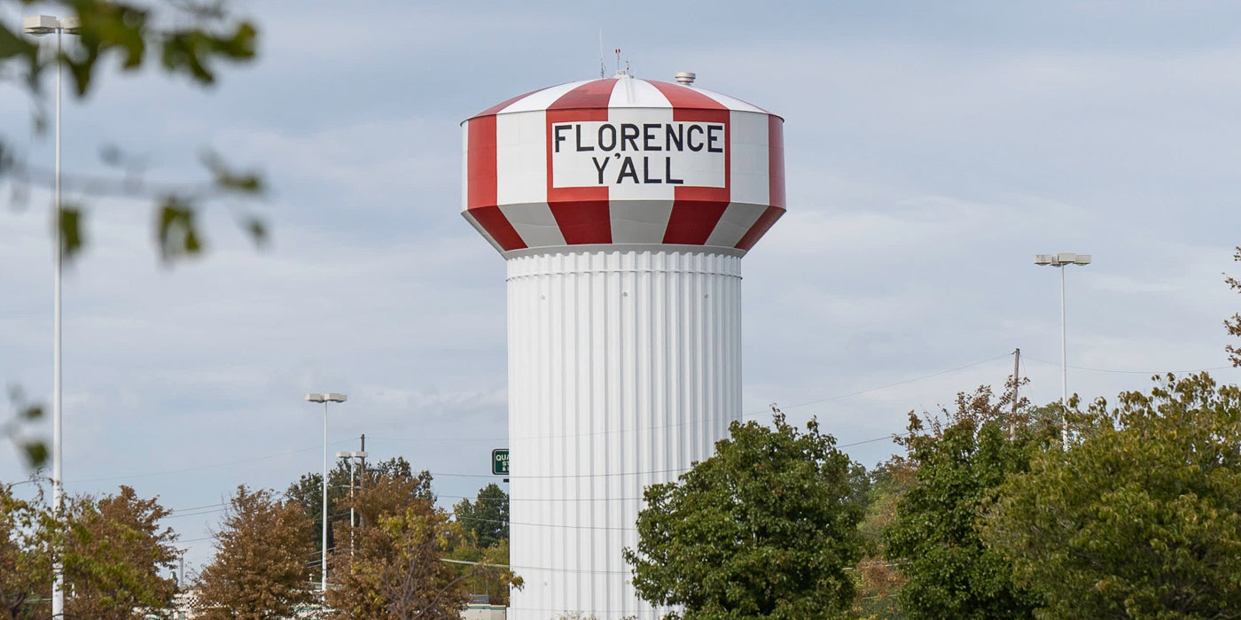 A large water tower in Florence, Kentucky, painted with red and white vertical stripes around the top and the words "FLORENCE Y'ALL" in bold black letters. The base of the tower is white and ribbed. Trees and streetlights are visible in the foreground under a cloudy sky.