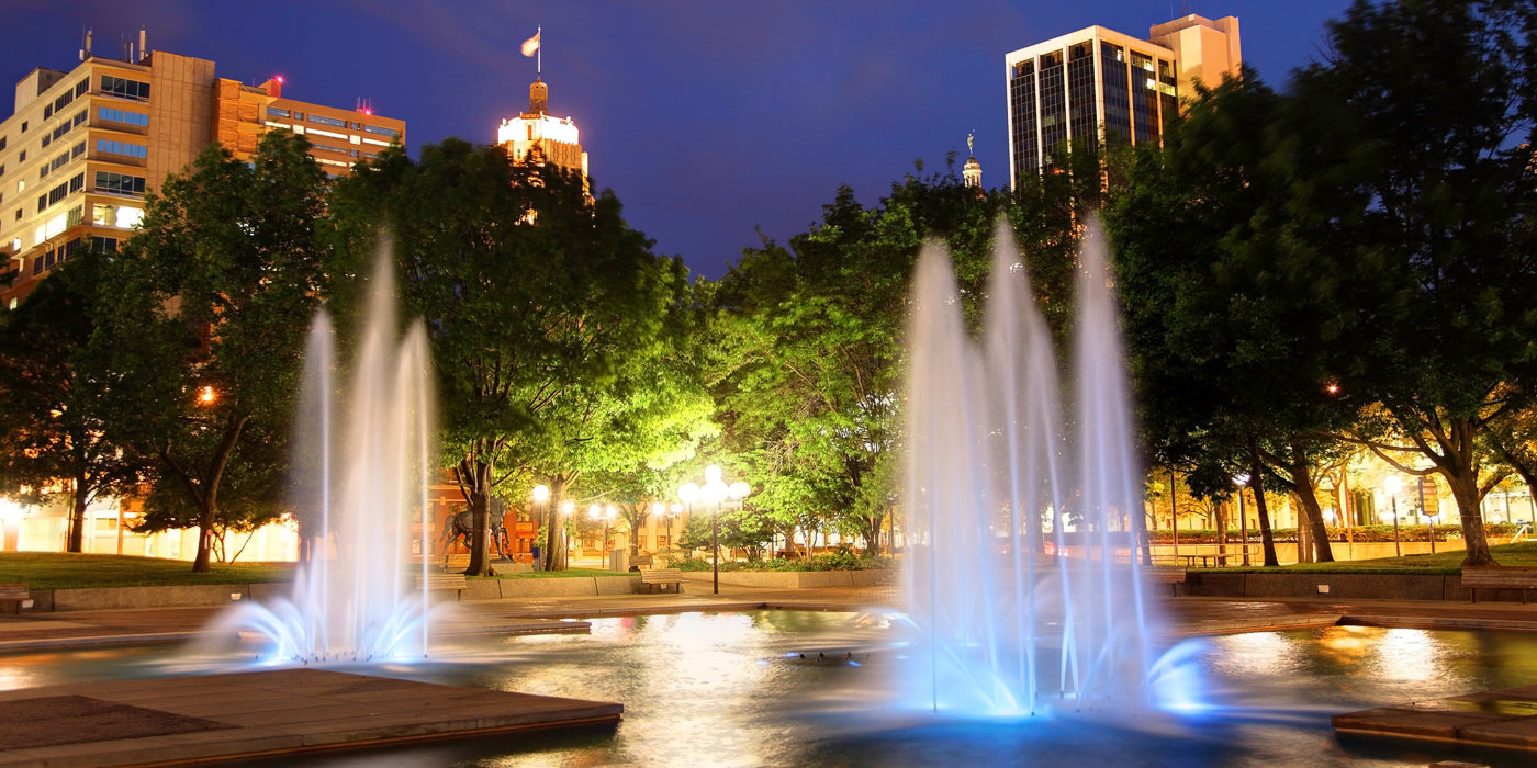 Nighttime view of a fountain in a city park in Fort Wayne, Indiana. The water jets are lit with blue lights, creating a vibrant glow. Trees surround the fountain, and tall buildings with illuminated windows rise in the background. The scene is calm and well-lit, with lamp posts casting a warm light on the greenery.