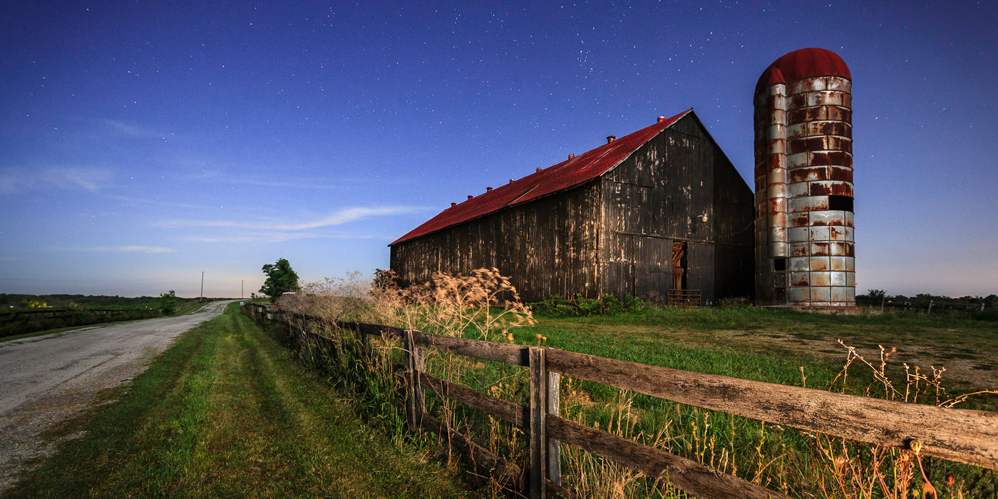 A nighttime rural scene in Georgetown, Kentucky, featuring an old wooden barn with a weathered red roof and a rusted metal silo beside it. The barn sits next to a narrow country road bordered by a wooden fence and tall grass. Stars are visible in the clear night sky above the peaceful countryside.