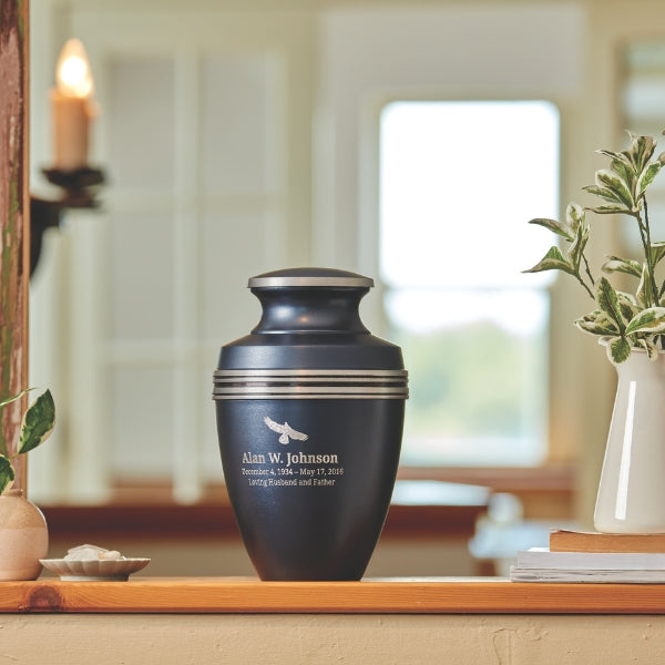 A matte blue urn with silver accent bands and a matching lid, displayed on a wooden ledge in a softly lit home setting. The urn features engraved text and a small emblem on the front, and engraving is available for personalization. A vase with greenery and stacked books are placed nearby.