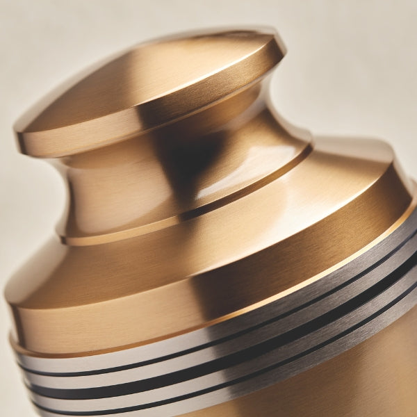 A close-up of a metallic gold urn with a polished finish. The image focuses on the urn’s domed lid and the decorative black and silver accent bands around its upper section. The reflective surface highlights the smooth craftsmanship.