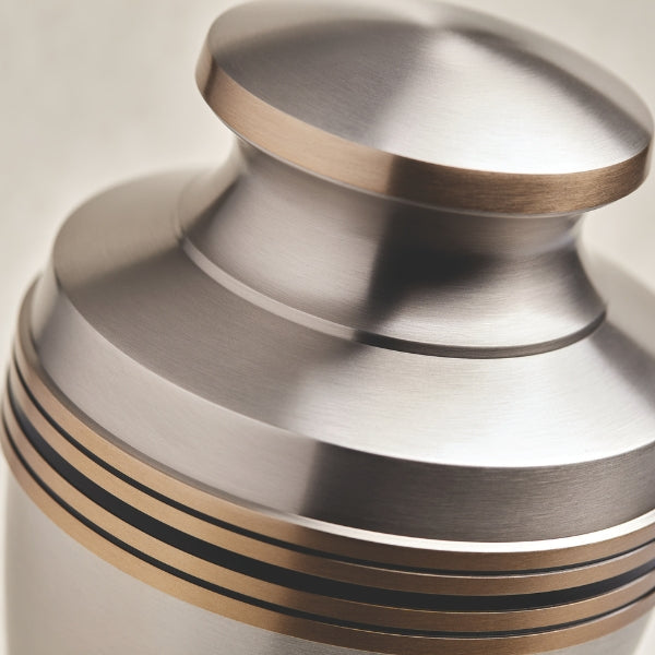 Close-up of a high-quality pewter memorial urn, showcasing its brushed silver finish with gold accent bands. The smooth, reflective surface and precision detailing highlight the urn’s craftsmanship, making it a sophisticated and timeless tribute.