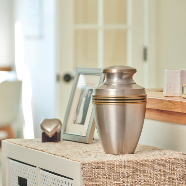 A brushed silver cremation urn with gold accent bands sits on a woven surface in a softly lit home setting. Beside it, a framed photograph and a heart-shaped keepsake create a touching tribute to a loved one. The urn’s timeless design and metallic sheen add a sense of elegance and reverence, making it a meaningful choice for honoring cherished memories.