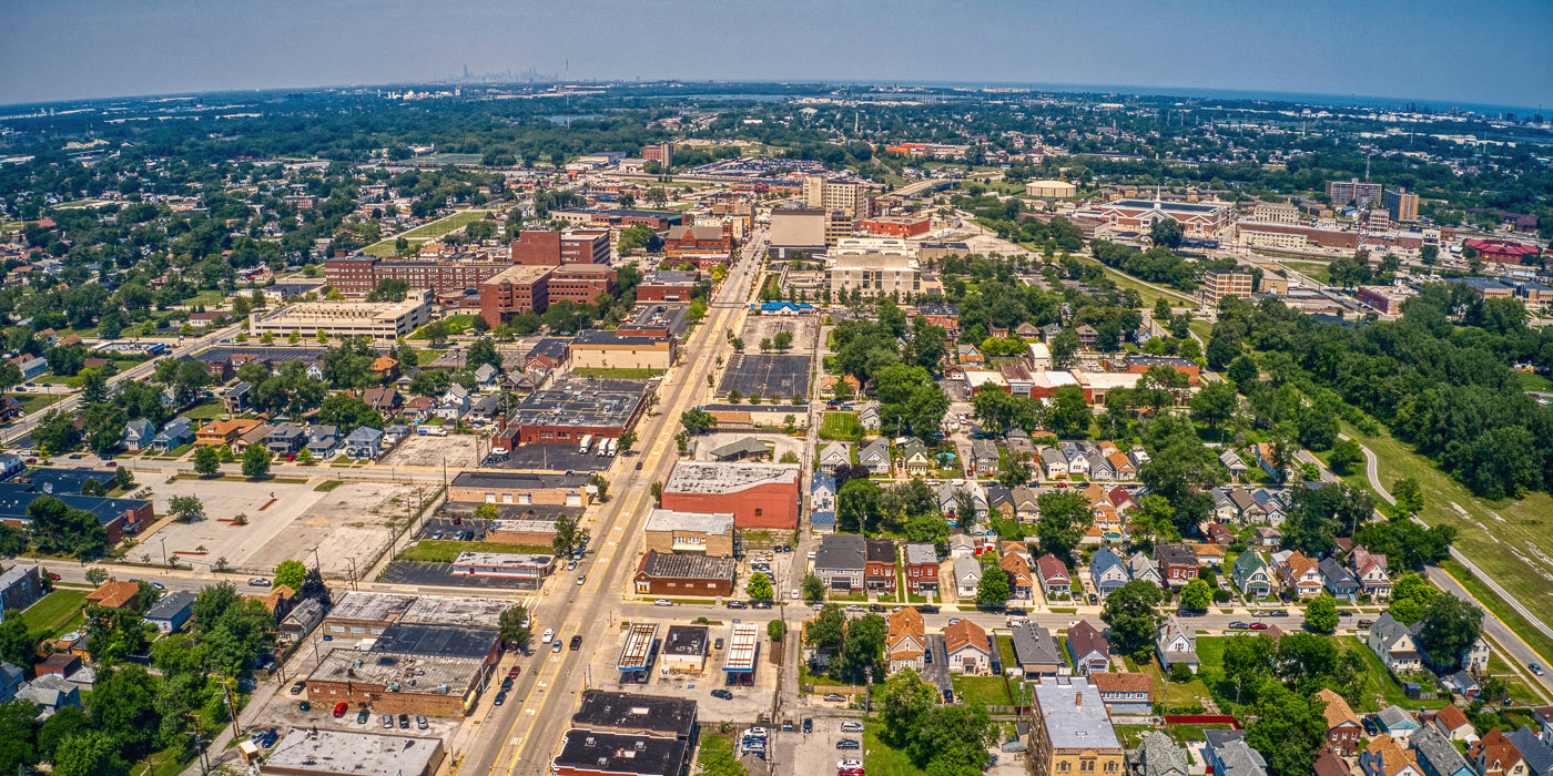 Aerial view of Hammond, Indiana, showing a mix of residential neighborhoods and commercial buildings. A main road runs vertically through the center, lined with businesses and parking lots. To the sides, tree-lined streets are filled with houses featuring various architectural styles. In the distance, larger institutional buildings and industrial facilities are visible under a hazy blue sky.