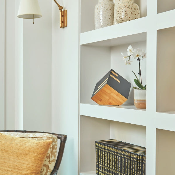 A modern cremation urn with a geometric cube design, featuring a matte black finish and natural bamboo accents. Displayed on a built-in shelf, the urn blends seamlessly with contemporary home decor, offering a discreet yet elegant tribute. Its unique tilted positioning adds an artistic touch, making it a meaningful and stylish memorial piece.