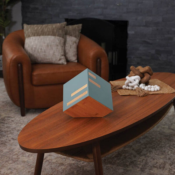 Modern cremation urn with sage green sides and bamboo accents displayed on a wooden coffee table in a stylish living room. The urn has a geometric cube shape with horizontal bamboo inlays. A brown leather chair with pillows and decorative objects are in the background.
