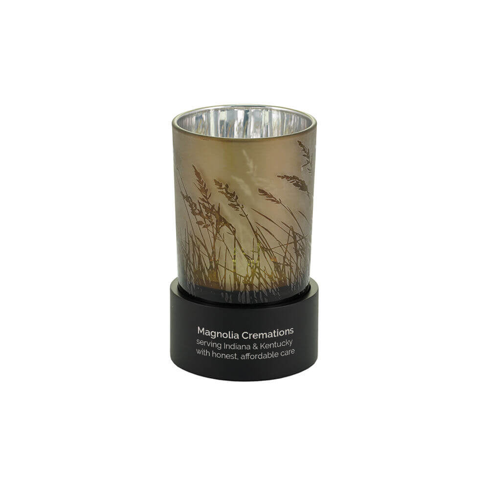 Keepsake urn that also functions as a tealight candle holder, featuring a frosted glass design with tall grasses and a black circular base. Optional engraving is shown.