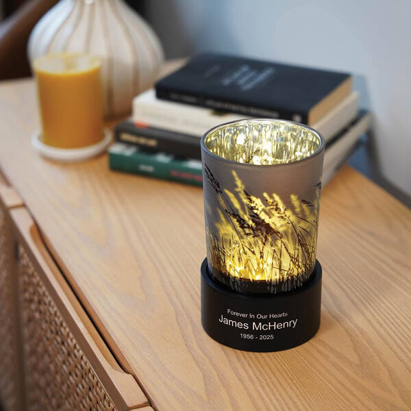 Keepsake urn that also serves as a tealight candle holder, shown lit on a wooden surface. The frosted glass features a design of tall grasses, and the black base displays optional engraving: "Forever In Our Hearts, James McHenry, 1956–2025." Books and a candle are visible in the background.