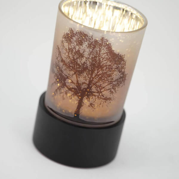 Close-up of a keepsake urn that also functions as a tealight candle holder. It features a frosted glass cup with a detailed tree silhouette design, softly illuminated from within, and sits on a black circular base.